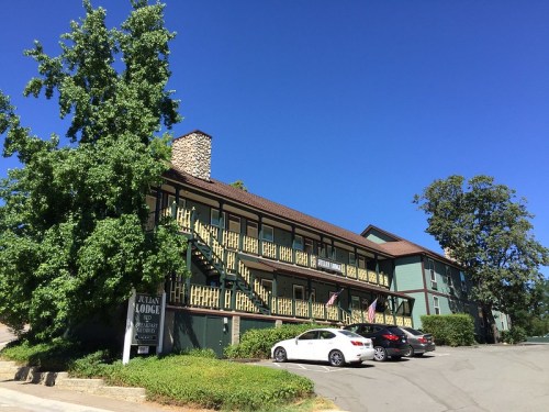 A green lodge with yellow accents, featuring a stone chimney and parked cars under a clear blue sky.