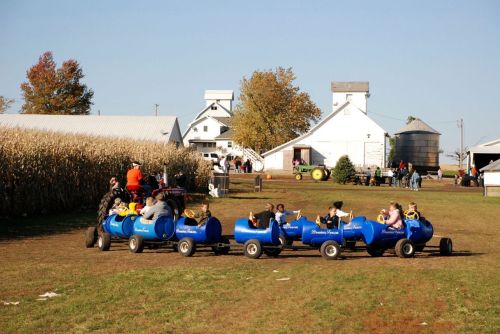 Children enjoy a ride on blue barrel-shaped carts at a farm, with barns and cornfields in the background.