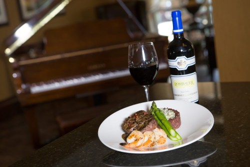 A gourmet meal featuring steak, shrimp, and asparagus, paired with a glass of red wine and a bottle, beside a piano.