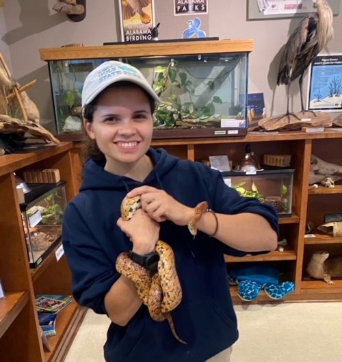 A smiling person holds a snake in a nature center, surrounded by displays of animals and plants.