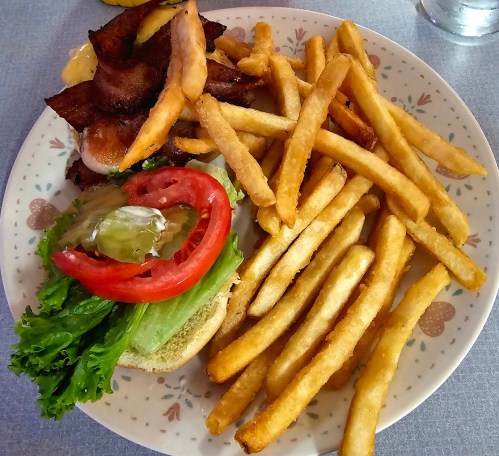 A plate with crispy French fries, a burger with lettuce, tomato, pickles, and bacon on a bun.