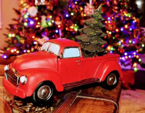 A red vintage truck with a Christmas tree in the back, set against a colorful, decorated Christmas tree backdrop.
