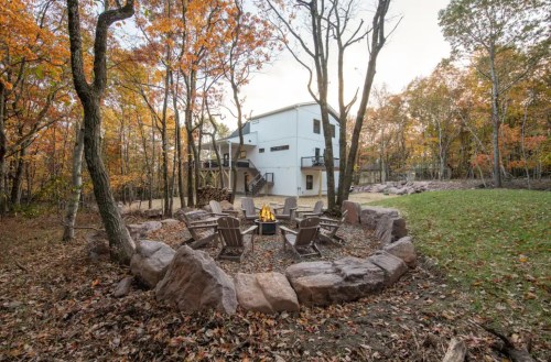 A cozy fire pit surrounded by chairs, with a modern house and autumn trees in the background.
