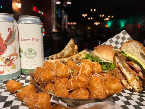 A plate of crispy tater tots, sandwiches, and two cans of beer on a checkered tablecloth in a dimly lit bar.