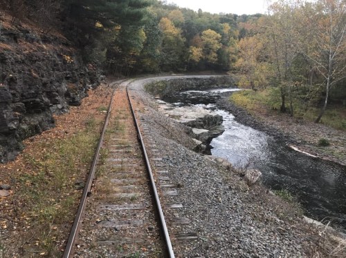 A winding railway track beside a river, surrounded by autumn foliage and rocky terrain.