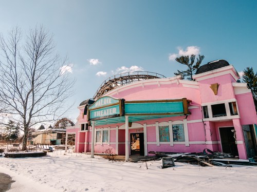 Abandoned pink theater building with a roller coaster in the background, surrounded by snow and bare trees.