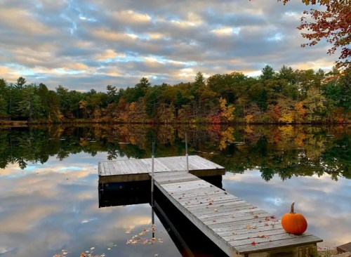 A serene lake scene with a wooden dock, colorful autumn trees, and a pumpkin resting on the edge.