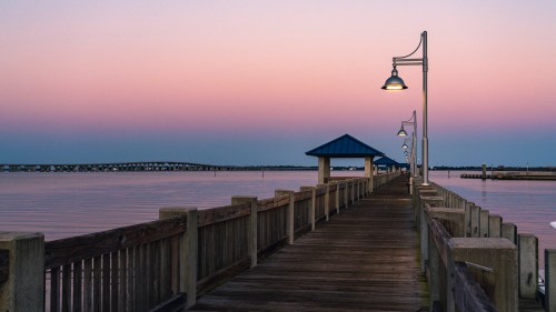 A wooden pier extends into calm waters at sunset, with lamps illuminating the path and a blue-roofed shelter in the distance.