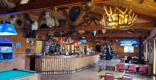 Interior of a rustic bar with wooden decor, a pool table, and mounted animal heads on the walls.