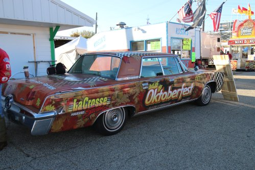 A vintage car wrapped in autumn leaves and Oktoberfest branding, parked at a festive outdoor event.