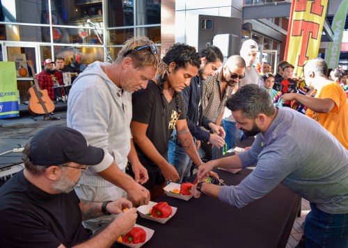 A group of men at a table preparing food during an outdoor event, with spectators in the background.