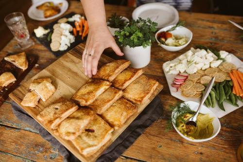 A hand reaches for a platter of golden pastries on a wooden table, surrounded by various appetizers and fresh vegetables.
