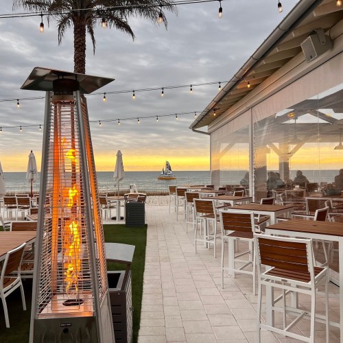 Cozy beachside restaurant with outdoor seating, a flame heater, and a sunset view over the ocean.