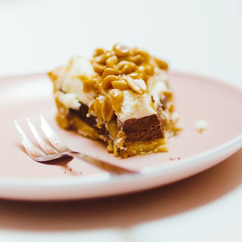 A slice of layered dessert with chocolate, cream, and nuts on a pink plate, accompanied by a fork.