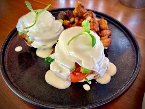 Two eggs Benedict on a black plate, topped with creamy sauce and garnished with microgreens, served with crispy potatoes.