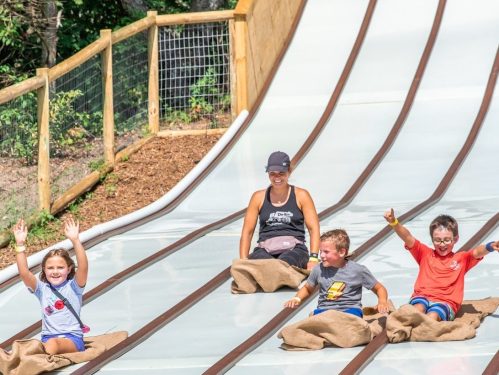 Four children and an adult slide down a water slide, smiling and having fun, with a wooden fence in the background.