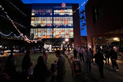 A lively outdoor event at dusk, with people mingling, colorful lanterns, and decorations around a building.