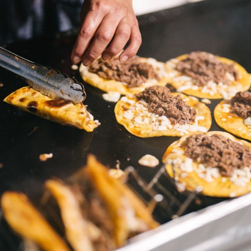 A hand using tongs to place beef-filled tortillas on a hot grill, with more tortillas cooking nearby.