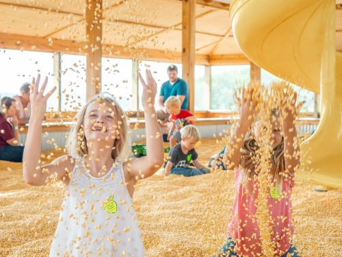 Two smiling girls play in a corn pit, tossing kernels in the air, with children and adults enjoying the background.