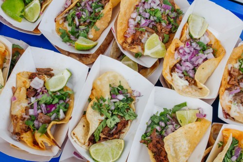 A colorful assortment of tacos topped with onions, cilantro, and lime wedges, served in paper trays.