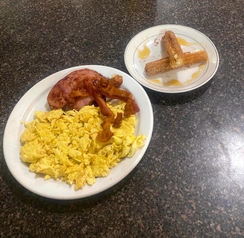 A plate with scrambled eggs, bacon, and a side of churros drizzled with syrup on a separate dish.