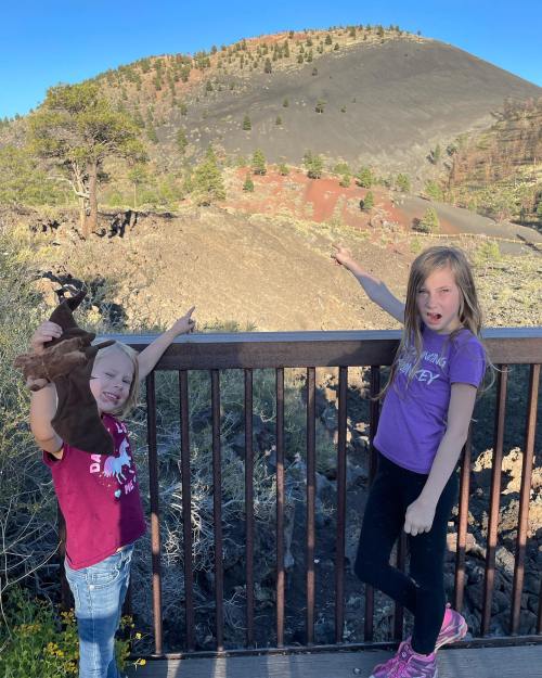 Two girls stand on a railing, pointing at a volcanic landscape with a mountain in the background.
