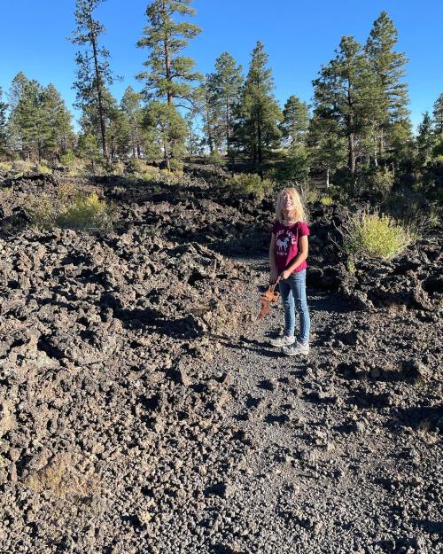 A child stands on a rocky path surrounded by trees and volcanic rock formations under a clear blue sky.