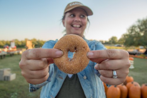 A smiling person holds a cinnamon sugar donut in front of a pumpkin patch on a sunny day.