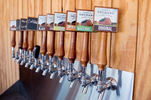 A row of cider taps with wooden handles and labeled signs, set against a wooden wall.