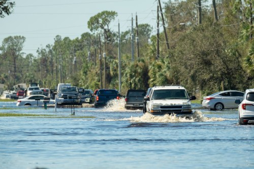 Vehicles navigate through a flooded street, with water covering the road and trees lining the background.
