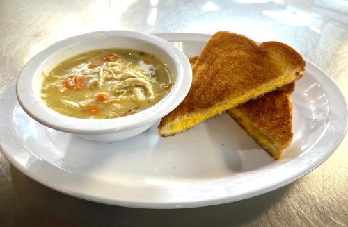 A bowl of chicken soup with vegetables next to two slices of grilled cheese on a white plate.
