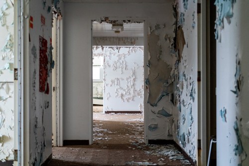A dimly lit hallway with peeling paint and debris scattered on the floor, leading to a doorway at the end.