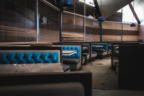 Abandoned diner interior with blue booths, empty tables, and dusty floors, featuring large windows and overhead lights.