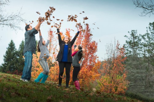 A family joyfully tosses autumn leaves in the air, surrounded by colorful fall foliage.