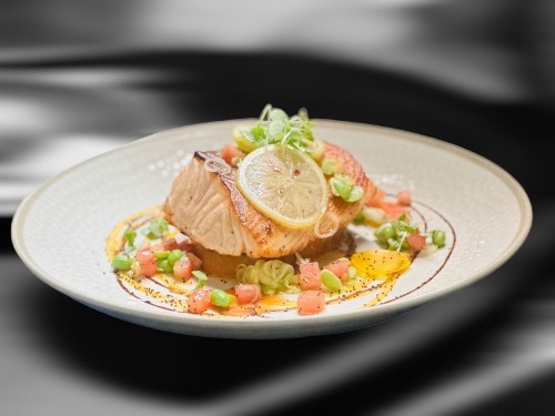 A beautifully plated salmon dish topped with lemon and garnished with green onions and diced tomatoes.