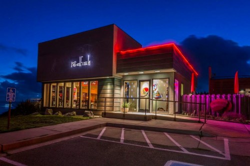 A modern restaurant exterior illuminated with colorful lights at dusk, featuring large windows and a welcoming entrance.