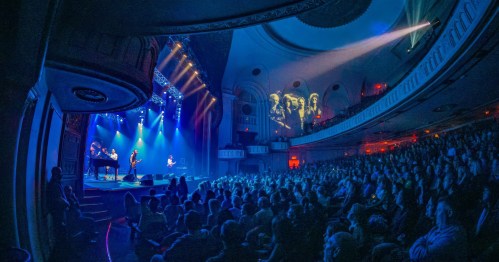 A lively concert scene with a band performing on stage, surrounded by a large audience in a beautifully lit theater.