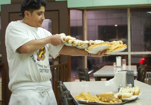 A man in a white shirt carries multiple plates of hot dogs and fries in a diner setting.
