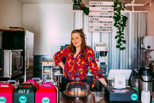 A smiling woman in a colorful shirt stands behind a counter in a cozy café, with menu items displayed above.