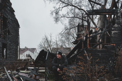 A person sits among the ruins of a burned building, surrounded by charred debris and overgrown vegetation.