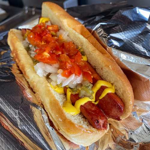 A close-up of a hot dog in a bun, topped with diced tomatoes, onions, mustard, and relish, wrapped in foil.