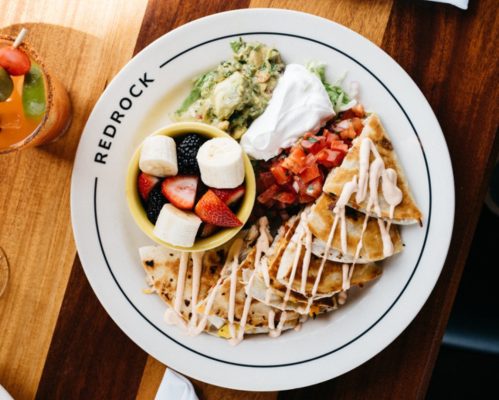 A plate of quesadillas drizzled with sauce, served with guacamole, salad, and a bowl of mixed fruit.