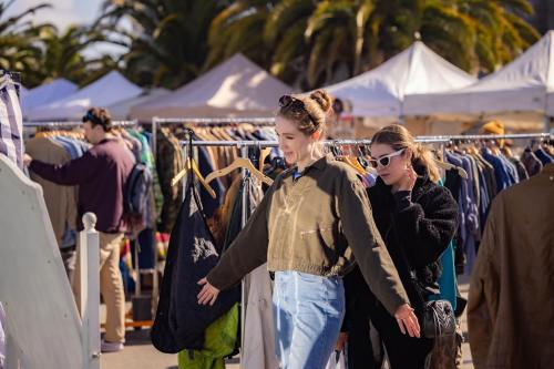 Two women browse clothing at an outdoor market, surrounded by racks of garments and palm trees in the background.