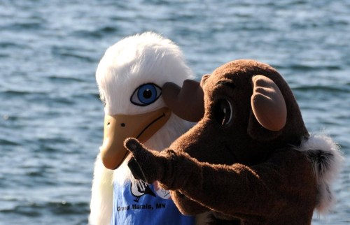 Two mascots stand by a body of water, one resembling a bird and the other a bison, both smiling and interacting.