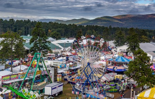 Aerial view of a vibrant fairground with rides, colorful tents, and mountains in the background under a cloudy sky.