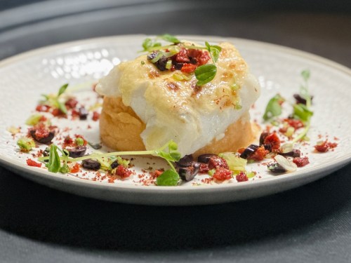 A beautifully plated dish featuring a piece of fish on a bed of soft bread, garnished with herbs and colorful toppings.