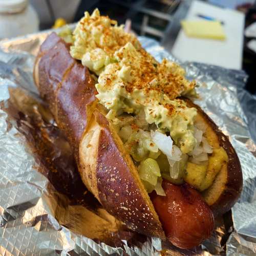 A gourmet hot dog in a pretzel bun, topped with mustard, onions, pickles, and a creamy spread, served on foil.