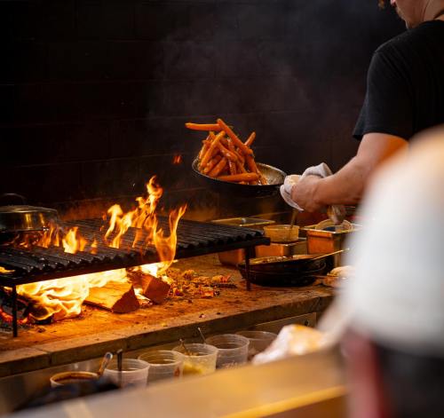 A chef tosses carrots in a pan over a grill with flames, surrounded by cooking utensils and containers.