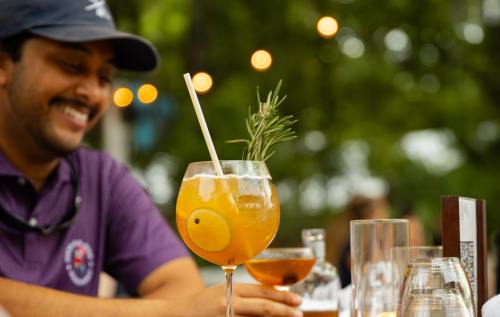 A man smiles while holding a cocktail with a lemon slice and rosemary, surrounded by a lively outdoor setting.