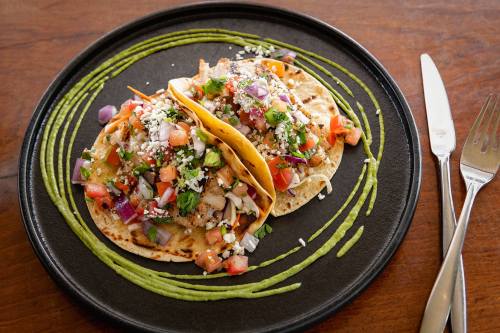 Two tacos filled with colorful toppings on a black plate, garnished with green sauce and fresh vegetables.